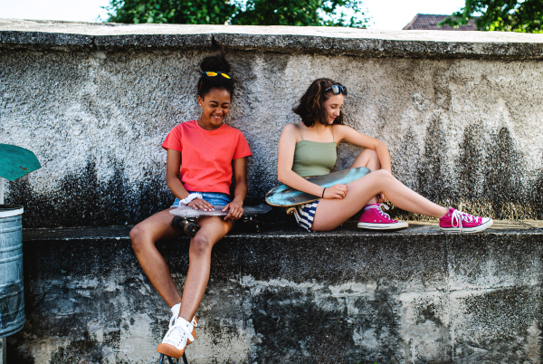 Young teenager girls friends with skateboards outdoors in city, sitting by concrete wall.