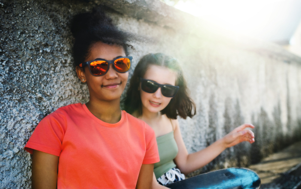 Young teenager girls friends with skateboards outdoors in city, sitting by concrete wall.