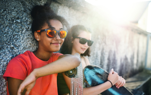 Young teenager girls friends with skateboards outdoors in city, sitting by concrete wall.