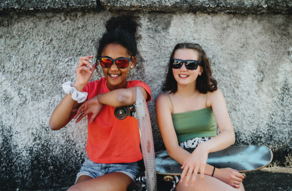 Young teenager girls friends with skateboards outdoors in city, sitting by concrete wall.