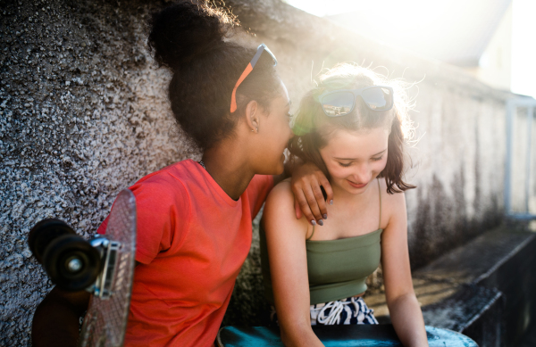 Young teenager girls friends with skateboards outdoors in city, sitting by concrete wall.