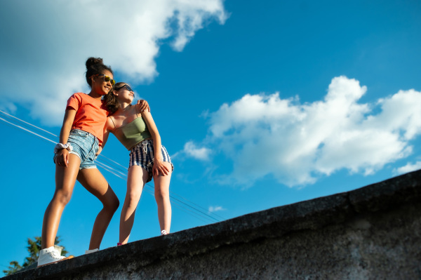 Young teenager girls friends with skateboards outdoors in city, sitting by concrete wall.