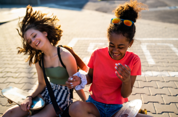 Teenager girls friends with skateboards sitting outdoors in city, having fun.