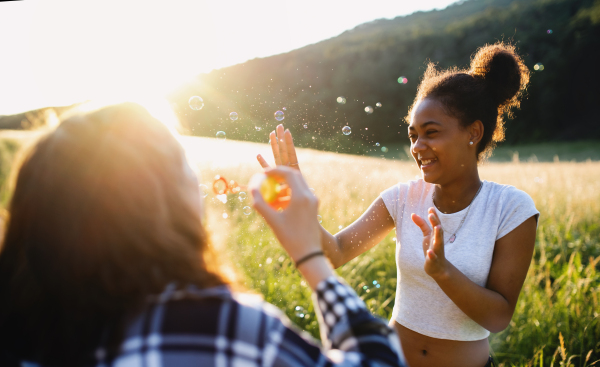 Cheerful young teenager girls friends outdoors in nature at sunset, blowing bubbles.