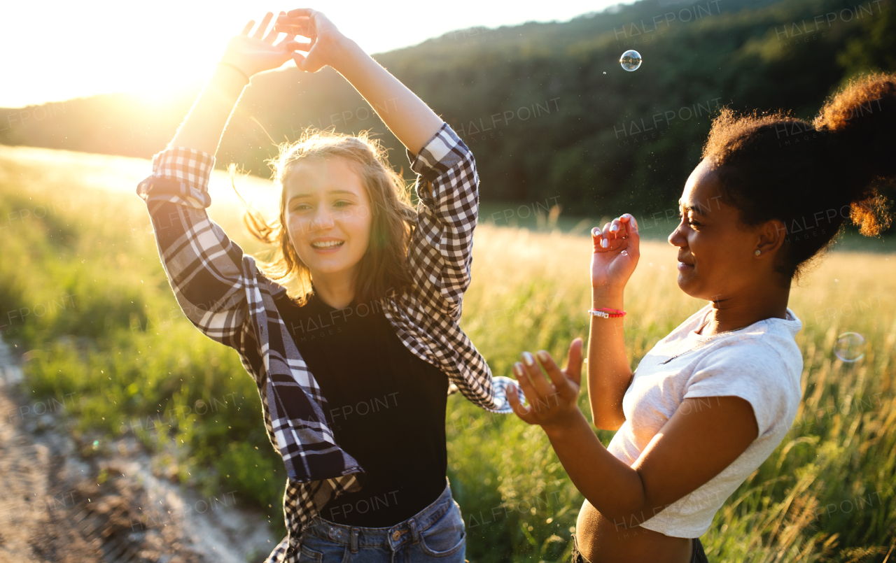 Cheerful young teenager girls friends outdoors in nature at sunset, blowing bubbles.