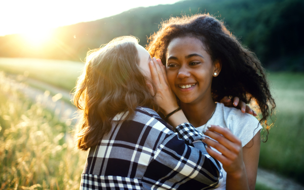 Cheerful young teenager girls friends outdoors in nature, whispering in ear.