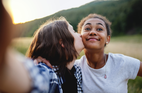 Front view of cheerful young teenager girls friends kissing outdoors in nature, taking selfie with smartphone.