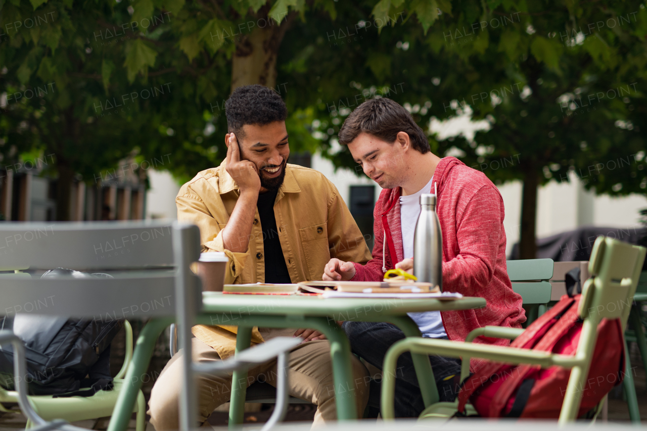 A young man with Down syndrome with his mentoring friend sitting outdoors in cafe and studying.