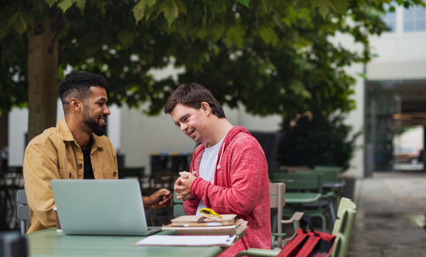 A young man with Down syndrome with his mentoring friend sitting outdoors in cafe using laptop.