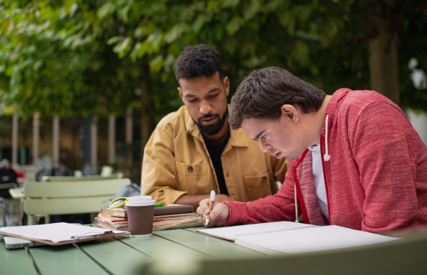 A young man with Down syndrome with his mentoring friend sitting outdoors in cafe and studying.