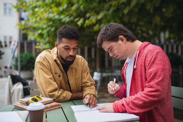 A young man with Down syndrome with his mentoring friend sitting outdoors in cafe and studying.