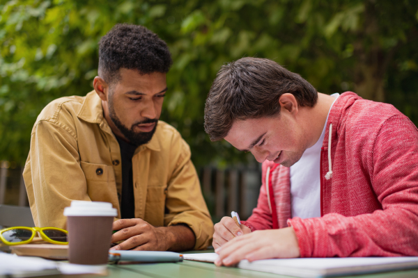 A young man with Down syndrome with his mentoring friend sitting outdoors in cafe and studying.