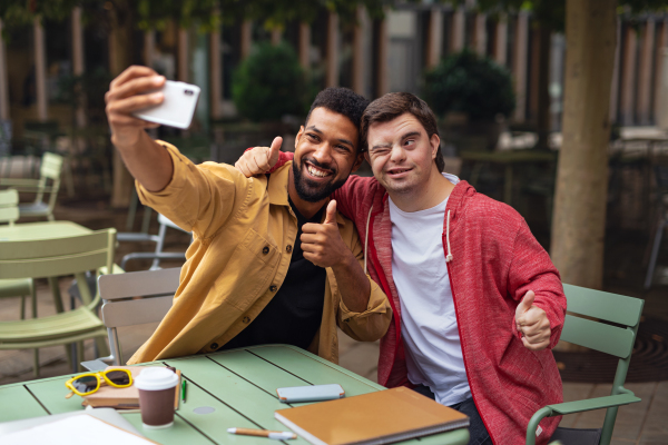 A young man with Down syndrome and his mentoring friend sitting and taking selfie outdoors in cafe