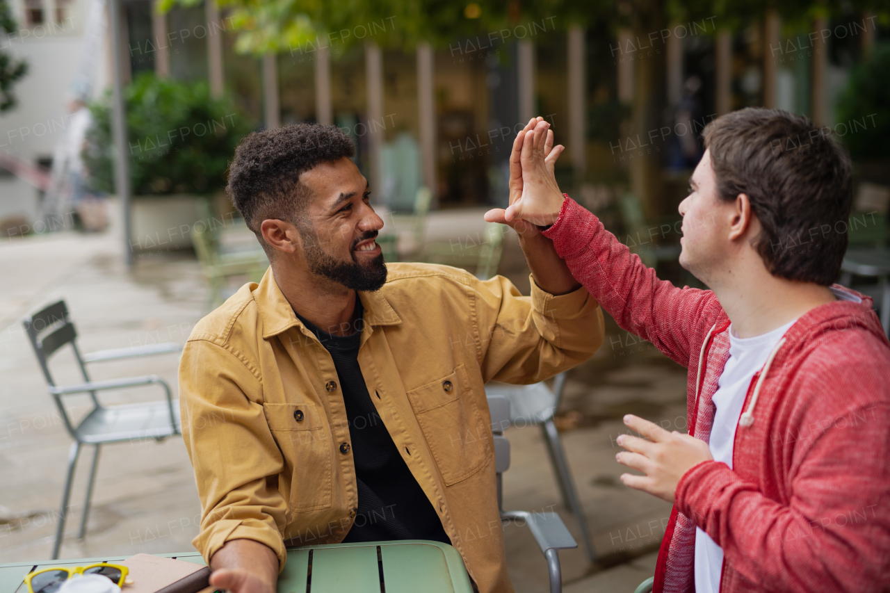 Young man with Down syndrome with his mentoring friend sitting outdoors in cafe high fiving.