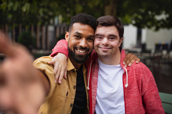 A young man with Down syndrome and his mentoring friend taking selfie nad lookig at camera outdoors