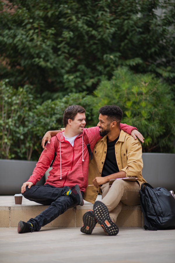 A happy young man with Down syndrome and mentoring friend sitting with arms around outdoors