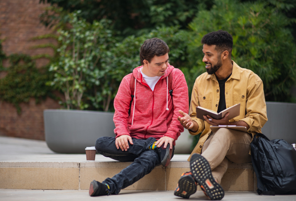 A happy young man with Down syndrome and mentoring friend sitting and talking outdoors