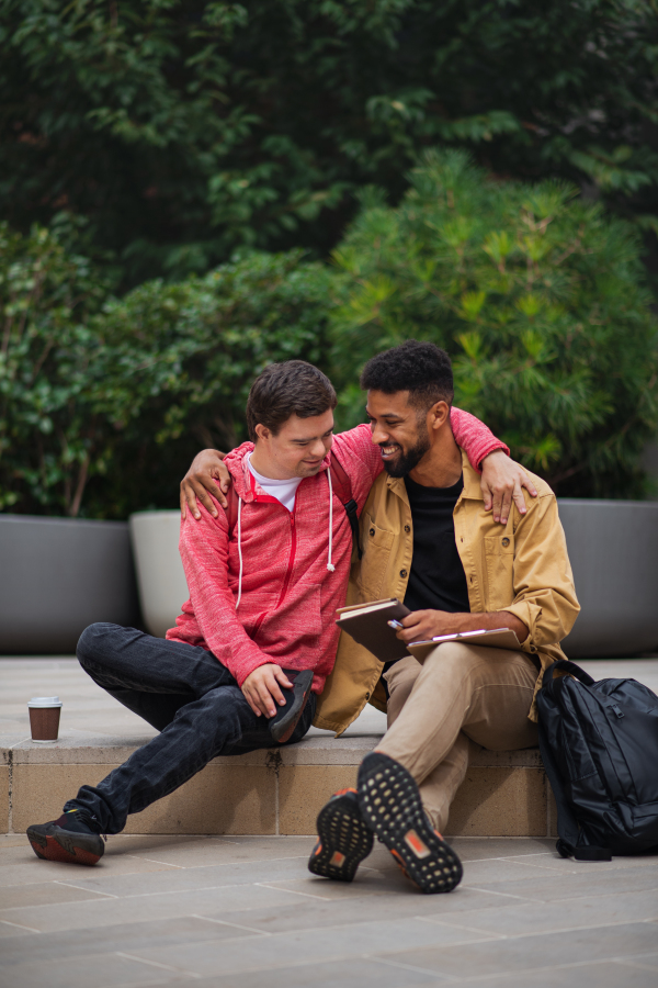 A happy young man with Down syndrome and mentoring friend sitting with arms around outdoors