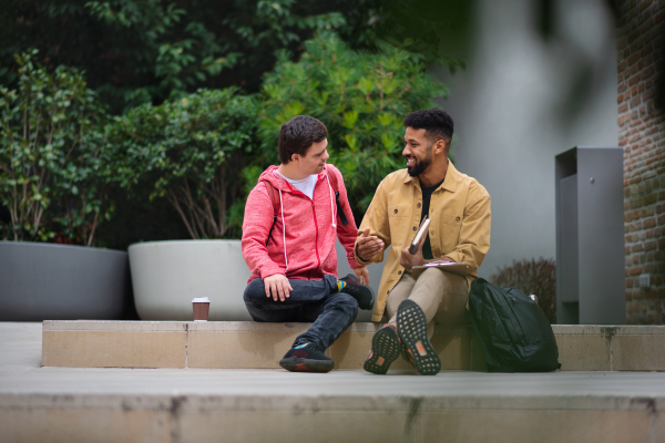 A happy young man with Down syndrome and mentoring friend sitting and talking outdoors