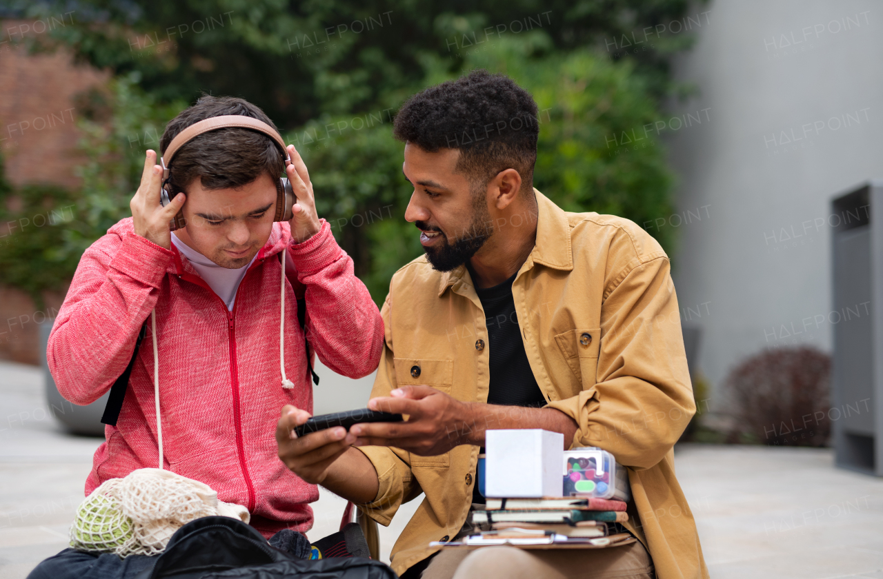 A young man with Down syndrome with his mentoring friend sitting outdoors and listening to music.