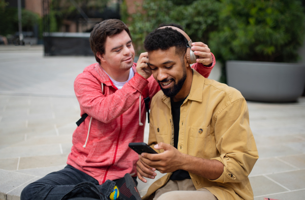 A young man with Down syndrome with his mentoring friend sitting outdoors and listening to music.