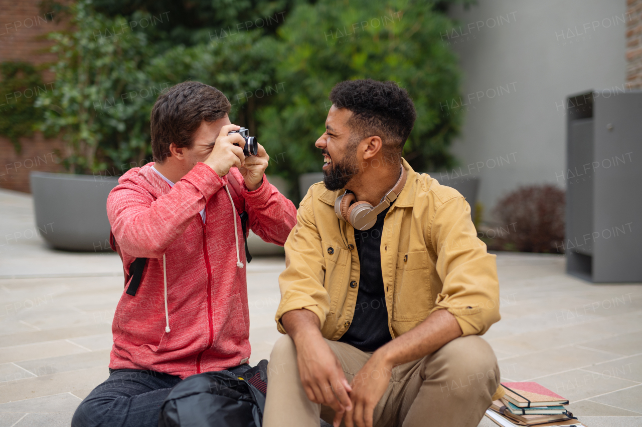 A happy young man with Down syndrome sitting and taking picture with camera of friend outdoors