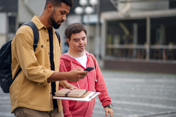 A young man with Down syndrome with his mentoring friend with smartphone walking and talking outdoors