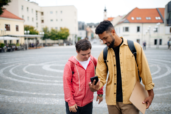 A young man with Down syndrome and his mentoring friend with smartphone walking and talking outdoors