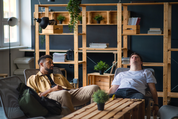 A young tired man with Down syndrome with his mentoring friend sitting indoors at school, taking break