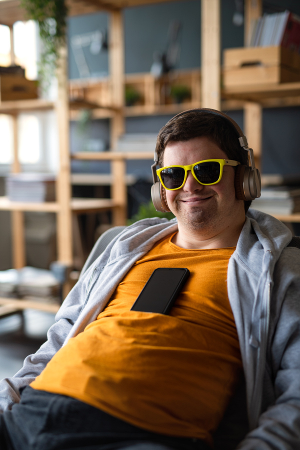 A relaxed young man with Down syndrome with sunglasses and headphones sitting indoors at school