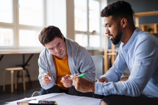 A young happy man with Down syndrome and his tutor studying indoors at school.