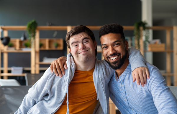 Happy young man with Down syndrome and his tutor with arms around looking at camera indoors at school