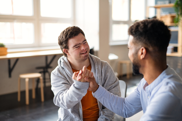 A young happy man with Down syndrome with his mentoring friend celebrating success indoors at school.