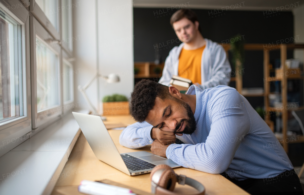 A tired young African-american teacher sleeping with head on desk indoors in staffroom.