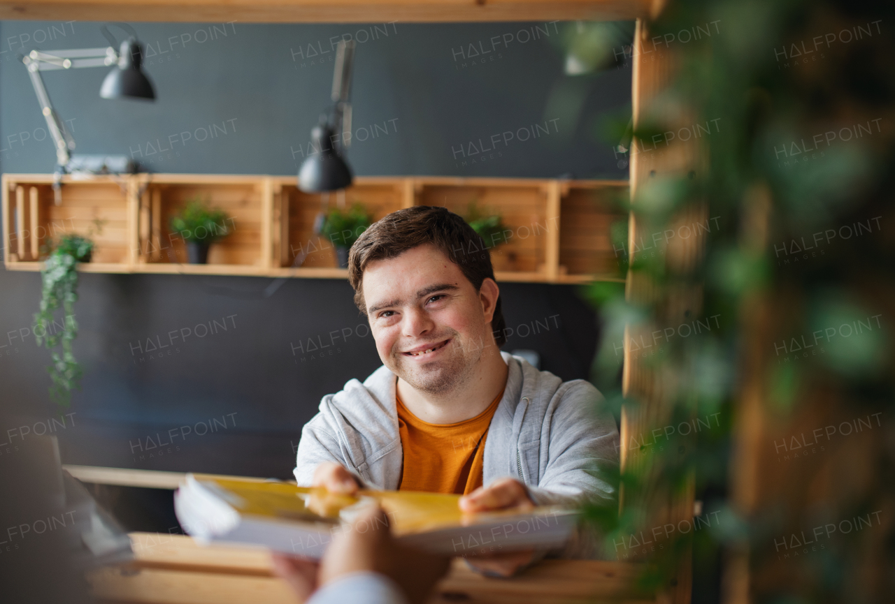 A happy young man with Down syndrome sitting and studying indoors at school, taking book from mentor