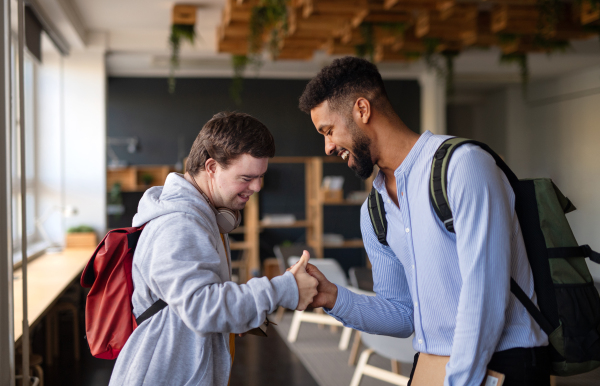 A young happy man with Down syndrome with his mentoring friend greeting indoors at school.