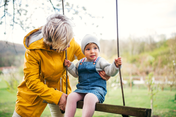 A senior grandmother with toddler granddaughter on a swing in garden in spring.