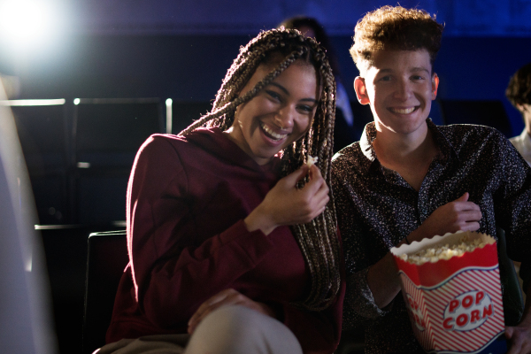 Cheerful young couple eating popcorn while watching movie in cinema, laughing.