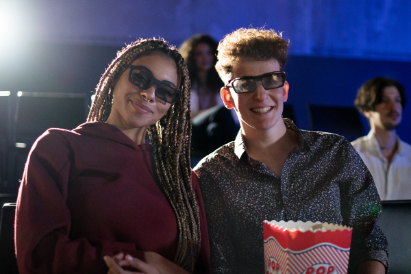 Young couple with 3d glasses and popcorn watching movie in cinema, looking at camera.