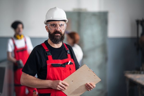 A portrait of worker with helmet and protective glasses indoors in factory looking at camera.