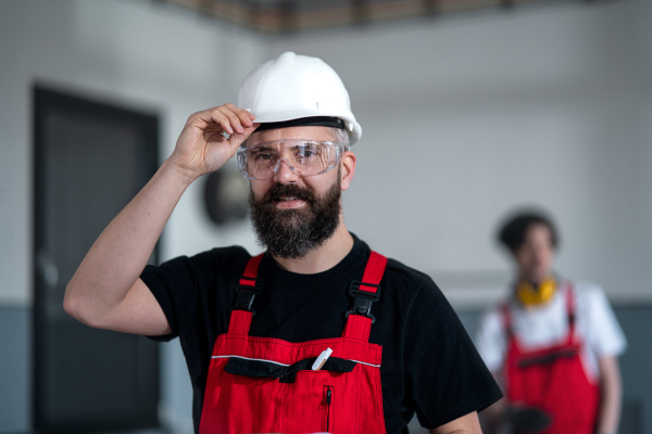 A portrait of worker with helmet and protective glasses indoors in factory looking at camera.