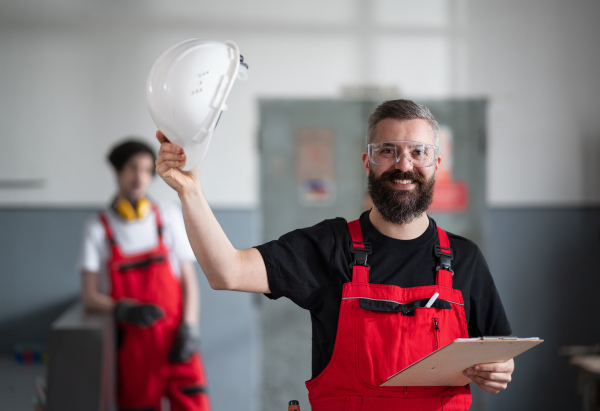 A portrait of cheerful worker with helmet and protective glasses indoors in factory looking at camera.