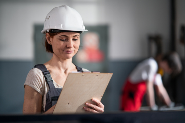 A low-angle view of worker with helmet and clipboard indoors in factory holding clipboard, working.