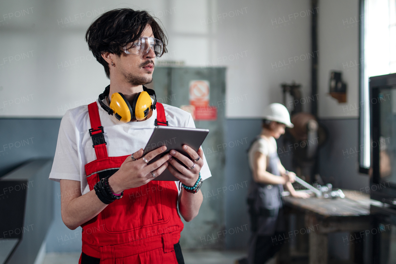 A portrait of young worker with helmet indoors in factory looking at camera.