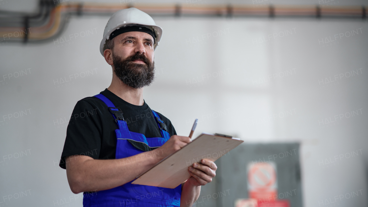 A low-angle view of worker with helmet indoors in factory holding clipboard.