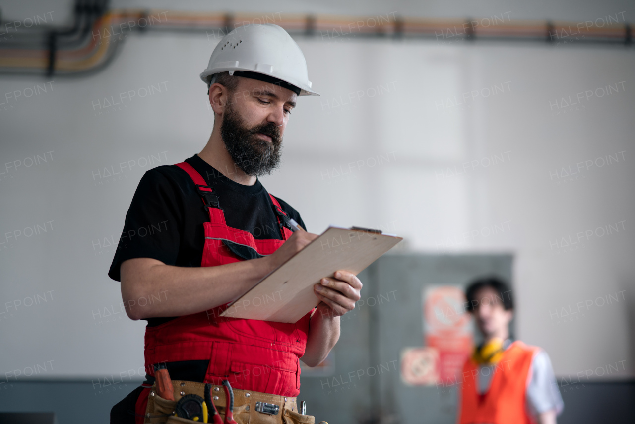 A man worker with helmet indoors in factory holding clipboard.