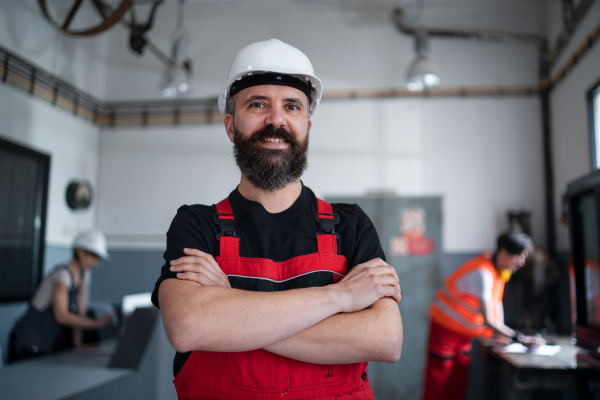 A portrait of worker with helmet indoors in factory looking at camera.