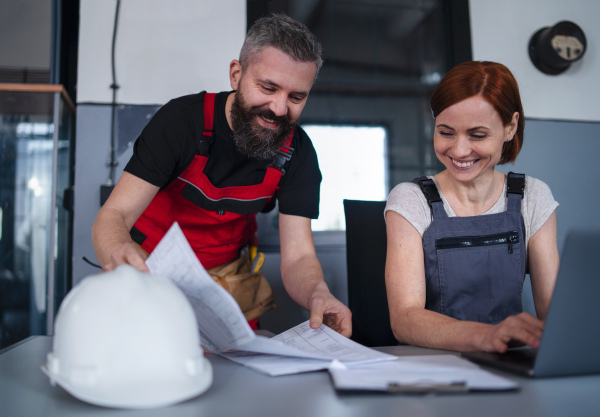 Mature man and woman workers or industry engineers with laptop indoors in factory, discussing issues.