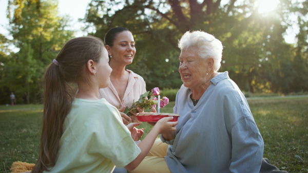 A small girl with mother giving cake to grandmother in a park, celebrating birthday.