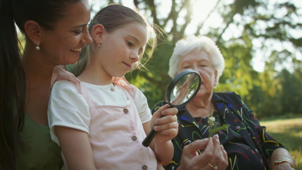 A small girl with mother and grandmother resting in a park, using magnifying glass to explore nature.